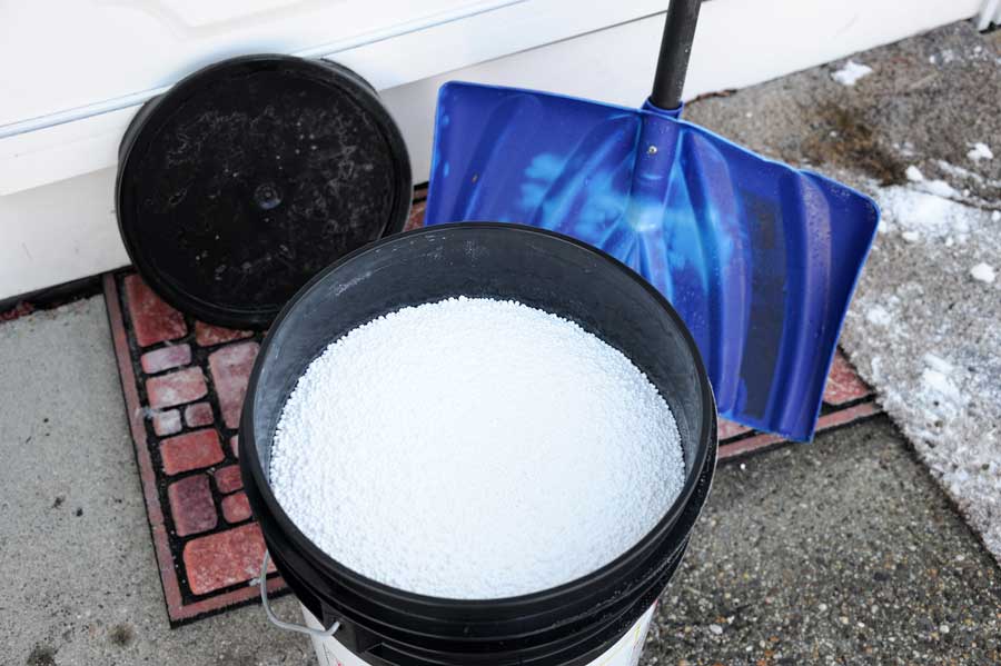 An open bucket of pavement salt and a snow shovel on a porch